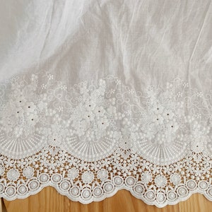 May include: White cotton fabric with a delicate white embroidered lace trim. The lace features a scalloped edge and a floral design.