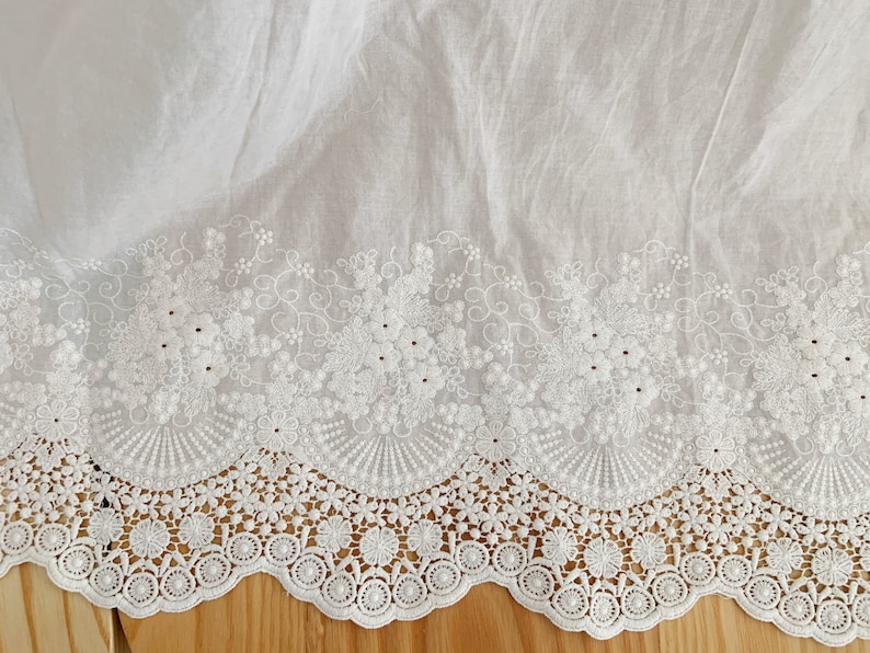 May include: White cotton fabric with a delicate white embroidered lace trim. The lace features a scalloped edge and a floral design.
