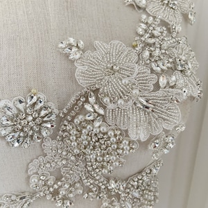 以下が含まれることがあります： 白い生地にビーズでできた花柄のデザインが施されています。デザインには白いパール、クリアなラインストーン、シルバーのビーズが使われています。
