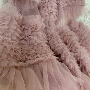 May include: Close-up of a dusty rose tulle fabric with multiple layers of ruffles. The fabric is soft and delicate, with a flowing texture.
