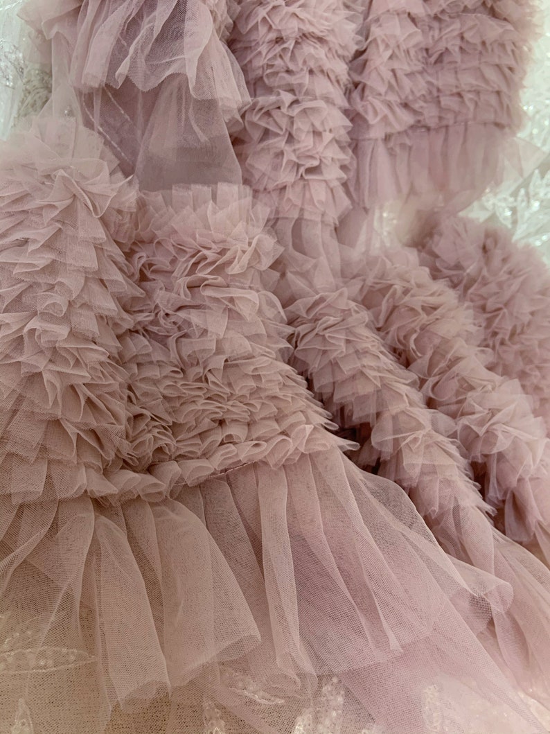 May include: Close-up of a dusty rose tulle fabric with multiple layers of ruffles. The fabric is soft and delicate, with a flowing texture.