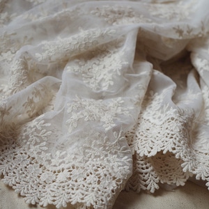May include: Ivory-colored lace fabric with floral embroidery. The delicate material is draped, showcasing the intricate details of the design. The edges feature a scalloped trim, adding a refined touch. Ideal for bridal wear or decorative projects.