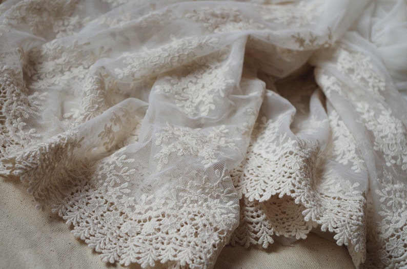 May include: Ivory-colored lace fabric with floral embroidery. The delicate material is draped, showcasing the intricate details of the design. The edges feature a scalloped trim, adding a refined touch. Ideal for bridal wear or decorative projects.