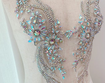 May include: Two symmetrical, iridescent rhinestone appliques. Each applique features floral and leaf designs with a central flower and trailing embellishments. The rhinestones are clear and reflect multiple colors. The appliques are on a white mesh backing.