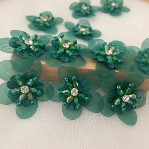 May include: Emerald green fabric flower embellishments with beaded centers and rhinestone accents. The flowers have layered petals and are arranged on a wooden surface. These decorative flowers are suitable for various craft projects.