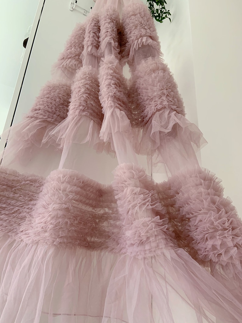 May include: A soft, pink tulle fabric with multiple layers of ruffles. The fabric is sheer and delicate, with a flowing, romantic look.