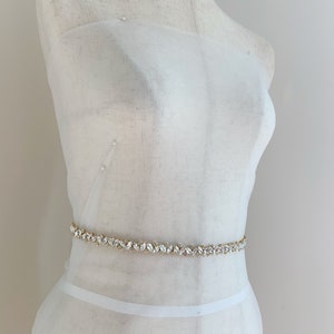Puede incluir: Una faja de tela blanca con un cintur&oacute;n adornado con cristales y dorado.