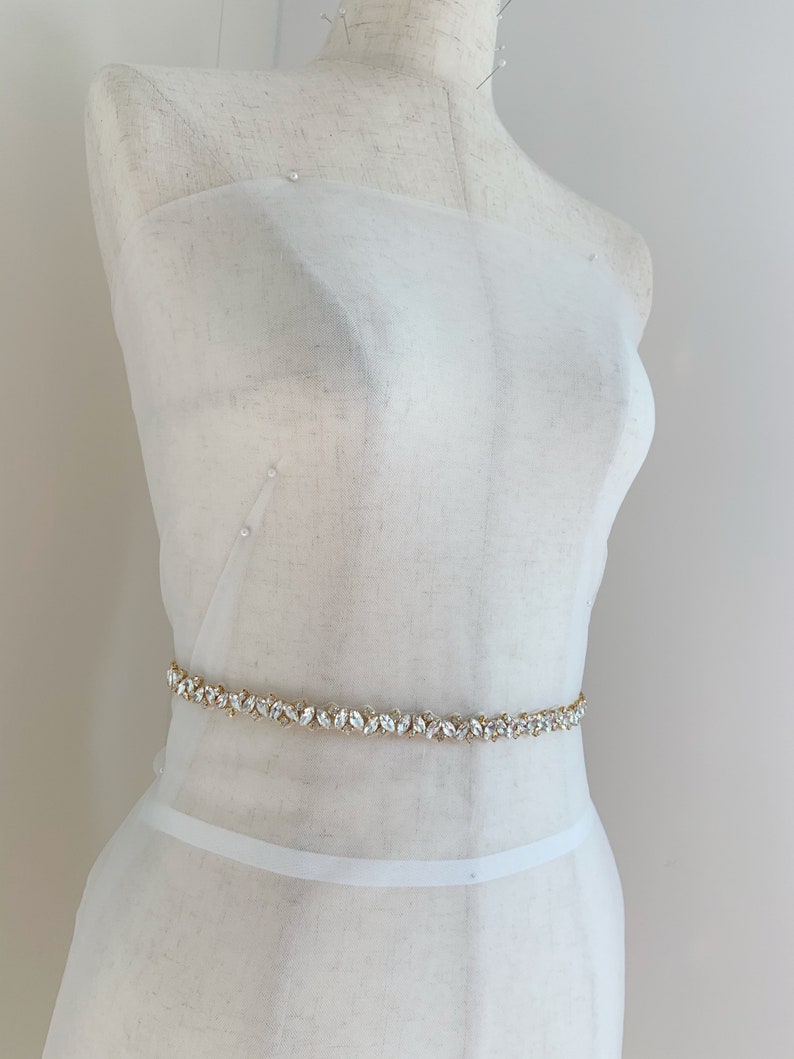 Puede incluir: Una faja de tela blanca con un cintur&oacute;n adornado con cristales y dorado.