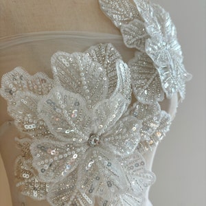 Pu&ograve; includere: Un'applicazione floreale bianca con paillettes e perle. L'applicazione &egrave; realizzata in tessuto delicato ed &egrave; perfetta per aggiungere un tocco di eleganza a qualsiasi capo.