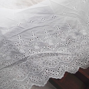 May include: White cotton fabric with a delicate eyelet lace trim. The lace features a repeating pattern of small flowers and leaves.