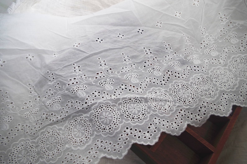 May include: White cotton fabric with a delicate eyelet lace trim. The lace features a repeating pattern of small flowers and leaves.