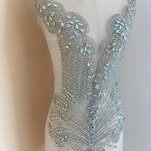 May include: A silver and iridescent rhinestone applique on sheer fabric. The design features a central, stylized feather shape with intricate detailing and a scalloped edge along the neckline and shoulders. The rhinestones vary in size and shape.