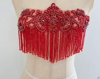 May include: A red beaded bodice with detailed floral and scrollwork patterns. The bodice has a fringe of hanging beads and is decorated with red gemstones. The garment is displayed on a mannequin.