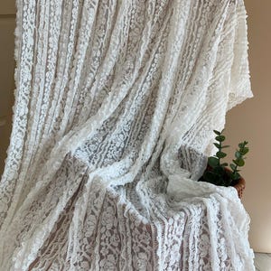 May include: White lace fabric with a floral design, draped over a surface. The lace has a delicate texture and is arranged in vertical stripes. A small basket with greenery is visible in the background.