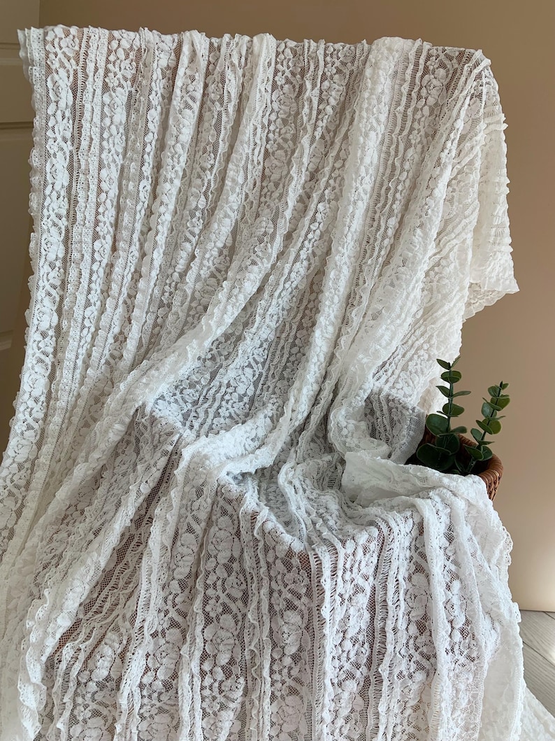 May include: White lace fabric with a floral design, draped over a surface. The lace has a delicate texture and is arranged in vertical stripes. A small basket with greenery is visible in the background.