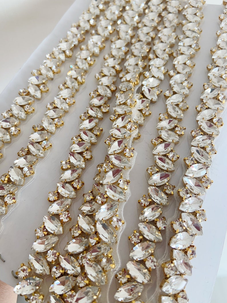 Puede incluir: Primer plano de un ribete de tela blanca con filas de cristales transparentes, facetados, en forma de marquesa, unidos a una cadena dorada.