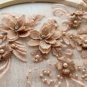 May include: A close-up of a delicate lace applique with a floral design. The applique is made of a soft, peach-coloured fabric and features intricate embroidery. The applique is adorned with small, pearl-like beads and sparkling rhinestones.