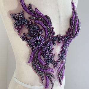 May include: A purple beaded applique with a floral design. The applique features various shades of purple beads and sequins, with silver accents. The design is on a sheer mesh backing, creating a delicate and ornate embellishment.