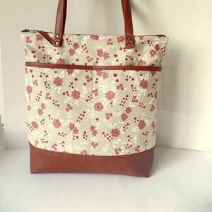 Op de afbeelding: Een bruine leren tote bag met een ritssluiting en een beige stoffen body met een bloemenpatroon met rode rozen en witte hartjes. De tas heeft een bruine leren bodem en twee bruine leren handvatten.