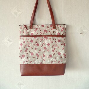 Op de afbeelding: Een bruine leren tote bag met een ritssluiting en een bloemenprint. De tas heeft een bruine leren bodem en twee bruine leren hengsels.