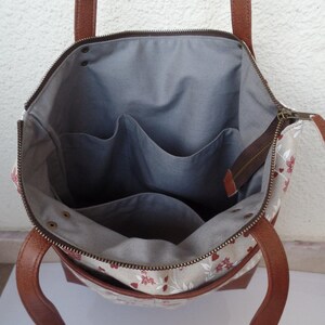 Op de afbeelding: Een beige canvas tote bag met bruine leren hengsels en een ritsvak. De tas heeft een bloemenpatroon in rood en wit en een grijze stoffen voering met drie binnenzakken.