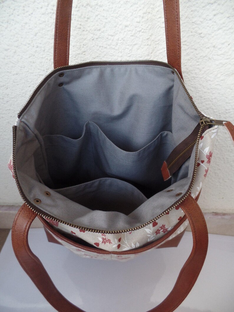 Op de afbeelding: Een beige canvas tote bag met bruine leren hengsels en een ritsvak. De tas heeft een bloemenpatroon in rood en wit en een grijze stoffen voering met drie binnenzakken.