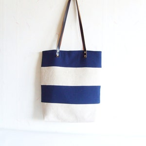May include: A striped canvas tote bag in navy blue and off-white, with brown leather handles secured by metal rivets. The bag is suspended against a white wall.