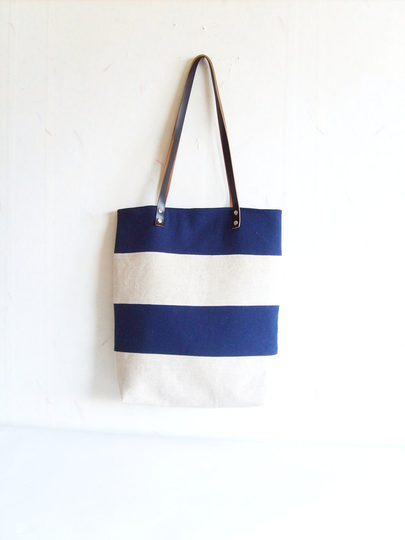 May include: A striped canvas tote bag in navy blue and off-white, with brown leather handles secured by metal rivets. The bag is suspended against a white wall.
