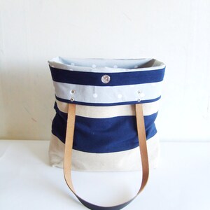 May include: A tote bag with a navy blue and beige striped design. It features light brown leather handles and a light grey interior with white polka dots. A silver snap closure is visible.