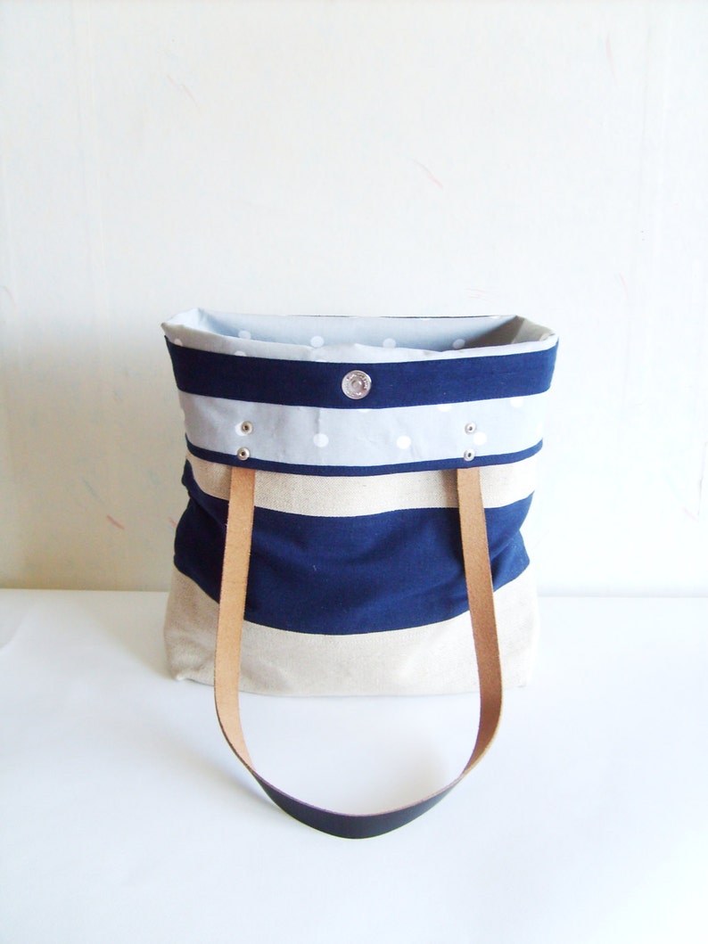 May include: A tote bag with a navy blue and beige striped design. It features light brown leather handles and a light grey interior with white polka dots. A silver snap closure is visible.