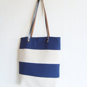 May include: A navy and cream striped tote bag with brown leather handles. The bag has a rectangular shape and a simple design. The handles are attached with metal rivets. Ideal for carrying items.
