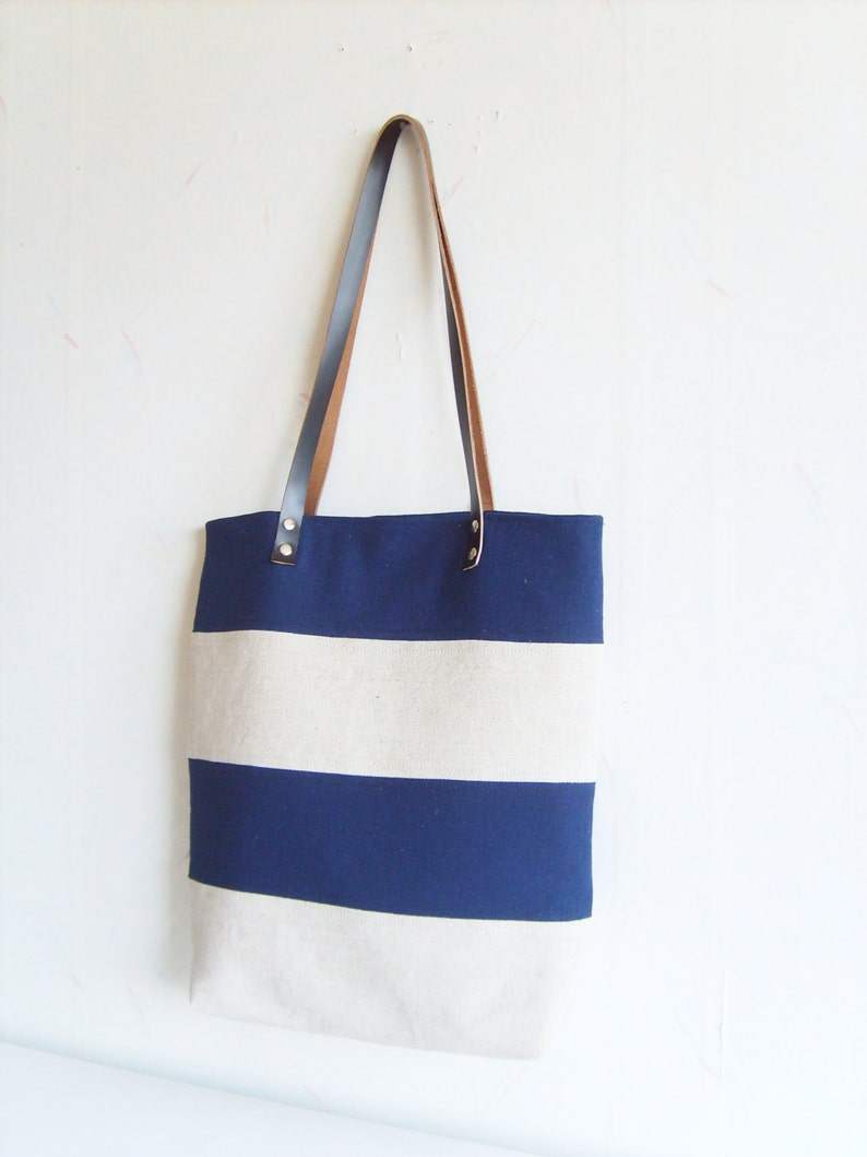 May include: A navy and cream striped tote bag with brown leather handles. The bag has a rectangular shape and a simple design. The handles are attached with metal rivets. Ideal for carrying items.