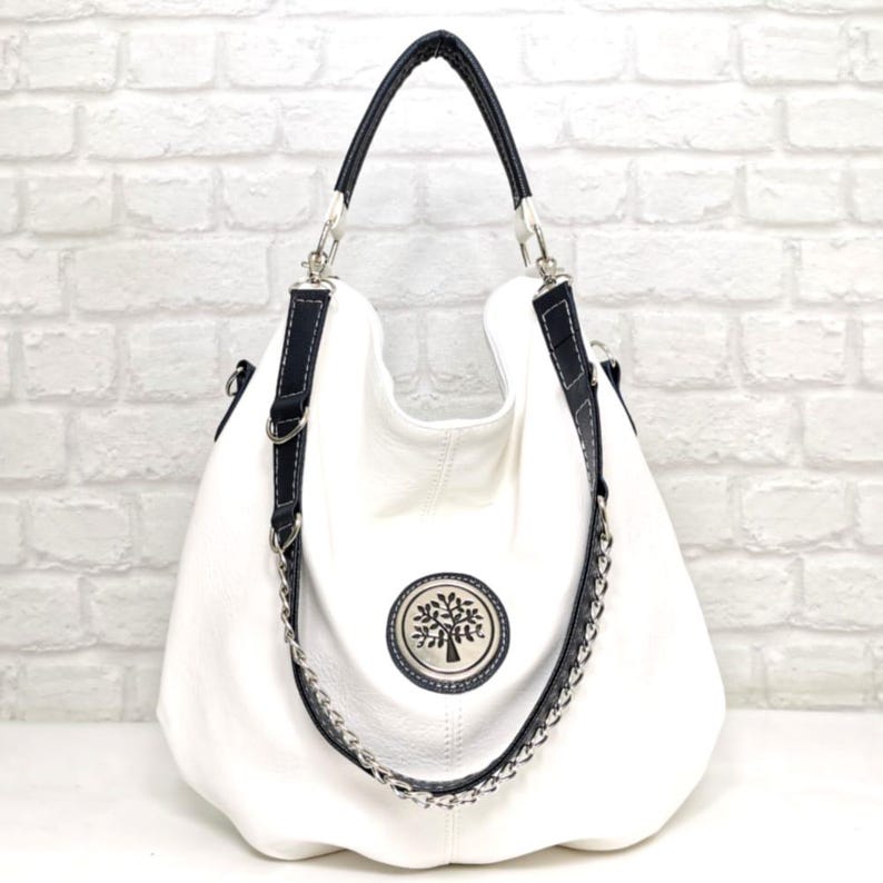 Peut inclure: Sac hobo en cuir blanc avec garnitures noires et d&eacute;tails argent&eacute;s. Il pr&eacute;sente une cha&icirc;ne argent&eacute;e et un embl&egrave;me circulaire avec un motif d'arbre. Le sac a une seule poign&eacute;e et une bandouli&egrave;re.