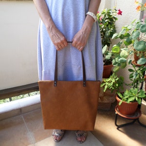 May include: A brown leather tote bag with brown leather straps. The bag is being held by a person wearing a light grey dress.