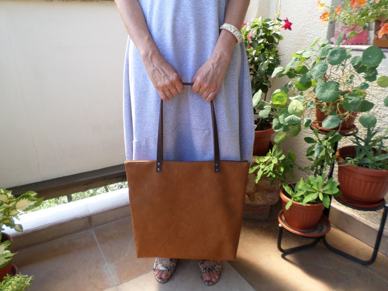 May include: A brown leather tote bag with brown leather straps. The bag is being held by a person wearing a light grey dress.