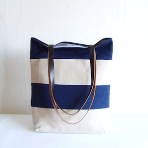 May include: A striped canvas tote bag in navy blue and beige, featuring brown leather handles. The bag's design includes horizontal stripes. It is a practical bag for carrying items, with a classic aesthetic.