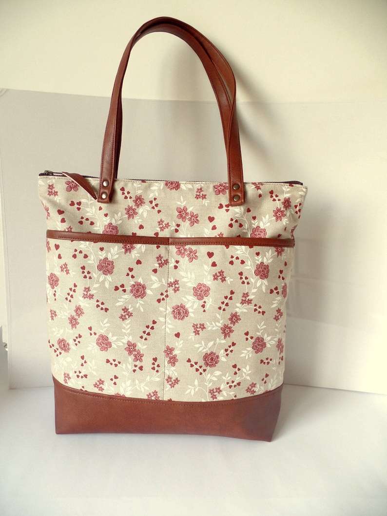 Op de afbeelding: Een bruine leren tote bag met een ritssluiting en een beige stoffen body met een bloemenpatroon met rode rozen en witte hartjes. De tas heeft een bruine leren bodem en twee bruine leren handvatten.