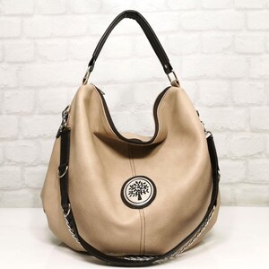 Peut inclure: Un sac hobo beige avec des bordures et des sangles noires. Le sac pr&eacute;sente un embl&egrave;me circulaire avec un motif d'arbre. Il poss&egrave;de une poign&eacute;e sup&eacute;rieure et une bandouli&egrave;re amovible avec une cha&icirc;ne argent&eacute;e. Le sac est pos&eacute; contre un mur de briques blanches.