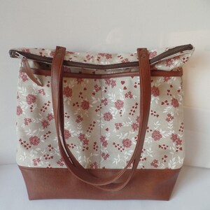 Op de afbeelding: Een grote bruine leren tote bag met een beige canvas body met een bloemenpatroon van rode rozen en witte hartjes. De tas heeft twee bruine leren hengsels en een ritssluiting.