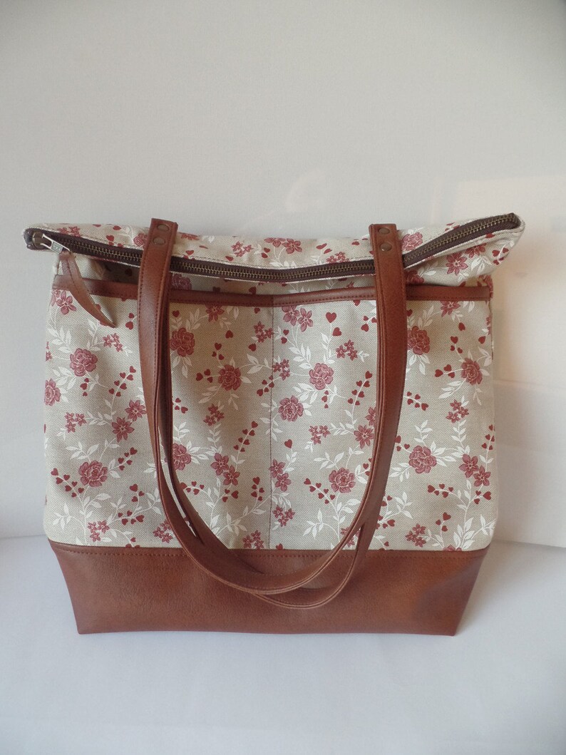 Op de afbeelding: Een grote bruine leren tote bag met een beige canvas body met een bloemenpatroon van rode rozen en witte hartjes. De tas heeft twee bruine leren hengsels en een ritssluiting.
