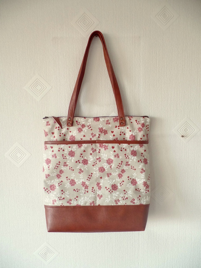 Op de afbeelding: Een bruine leren tote bag met een ritssluiting en een bloemenprint. De tas heeft een bruine leren bodem en twee bruine leren hengsels.