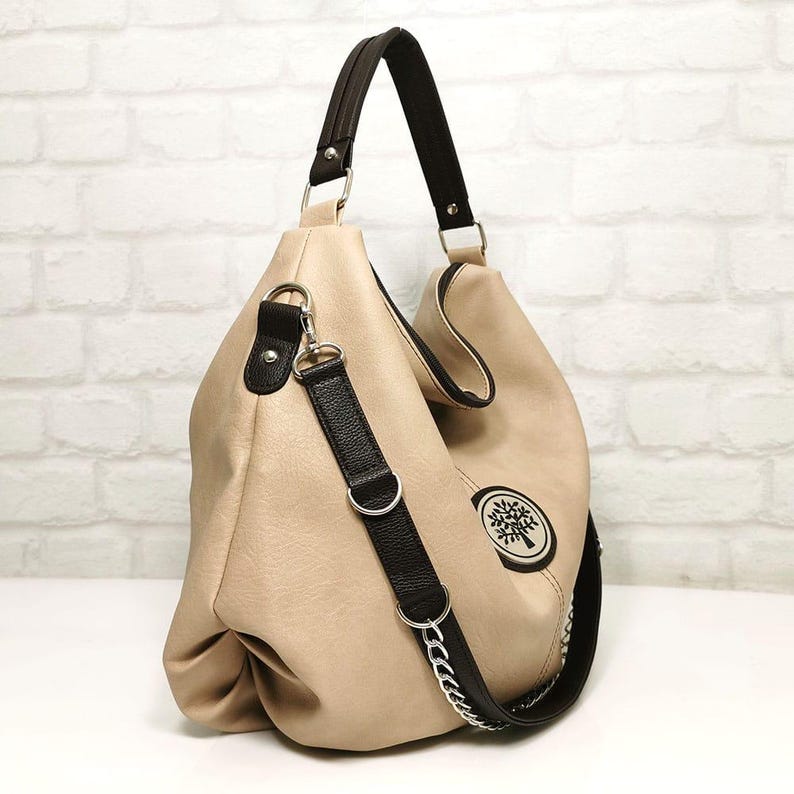 Peut inclure: Sac hobo beige avec une bandouli&egrave;re et une poign&eacute;e noires. Le sac pr&eacute;sente un logo d'arbre &agrave; l'avant et une cha&icirc;ne argent&eacute;e sur la bandouli&egrave;re. Le sac a un design souple et ample.