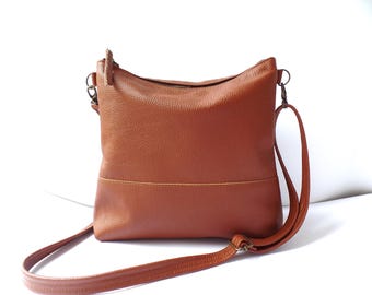 May include: A brown leather crossbody bag with a long adjustable strap. The bag has a rectangular shape with a zipper closure at the top. The bag is made of a textured leather material.