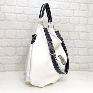 Peut inclure: Sac &agrave; main en cuir blanc avec des d&eacute;tails bleu marine. Il comporte une bandouli&egrave;re en cha&icirc;ne argent&eacute;e et un embl&egrave;me d&eacute;coratif. Le sac a une poign&eacute;e sup&eacute;rieure et une bandouli&egrave;re amovible. Sur un mur de briques blanches.