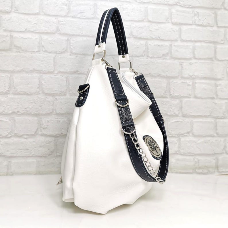 Peut inclure: Sac &agrave; main en cuir blanc avec des d&eacute;tails bleu marine. Il comporte une bandouli&egrave;re en cha&icirc;ne argent&eacute;e et un embl&egrave;me d&eacute;coratif. Le sac a une poign&eacute;e sup&eacute;rieure et une bandouli&egrave;re amovible. Sur un mur de briques blanches.