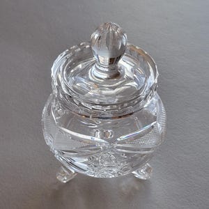 May include: A clear, cut-glass jar with a lid and three small feet. The jar has a round body with intricate geometric patterns and a decorative knob on top. The glass sparkles, reflecting light, and is suitable for storing small items.