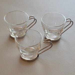 May include: Three clear glass espresso cups with silver-colored metal handles and bases. The cups have a simple, elegant design, perfect for serving coffee or other beverages. The cups are arranged on a light-colored surface.