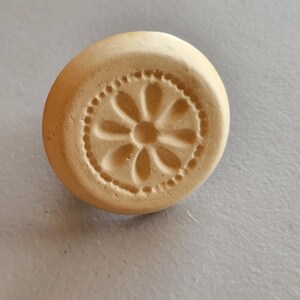 May include: A round, beige ceramic stamp with a floral design. The stamp features a central flower with petal shapes and a dotted border. The stamp is likely used for decorative purposes or crafting.