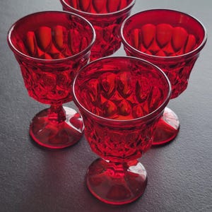 May include: Four ruby red goblets with a faceted design. The stemware has a round base and a short stem. The glasses are arranged on a dark surface, showcasing their vibrant color and intricate details. Ideal for serving beverages.