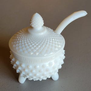 May include: A white, hobnail-style, lidded serving dish with a handle. The dish has a textured, geometric pattern on the lid and body, with a knob on top. It sits on three small feet. The handle curves upward.